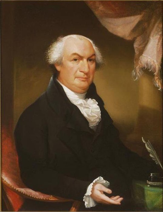 A formal portrait of an older man with thinning white hair, wearing a black coat and white ruffled shirt, seated with one hand on a green surface and a quill nearby—evoking the spirit of Constitution Day—against draped fabric in the background.