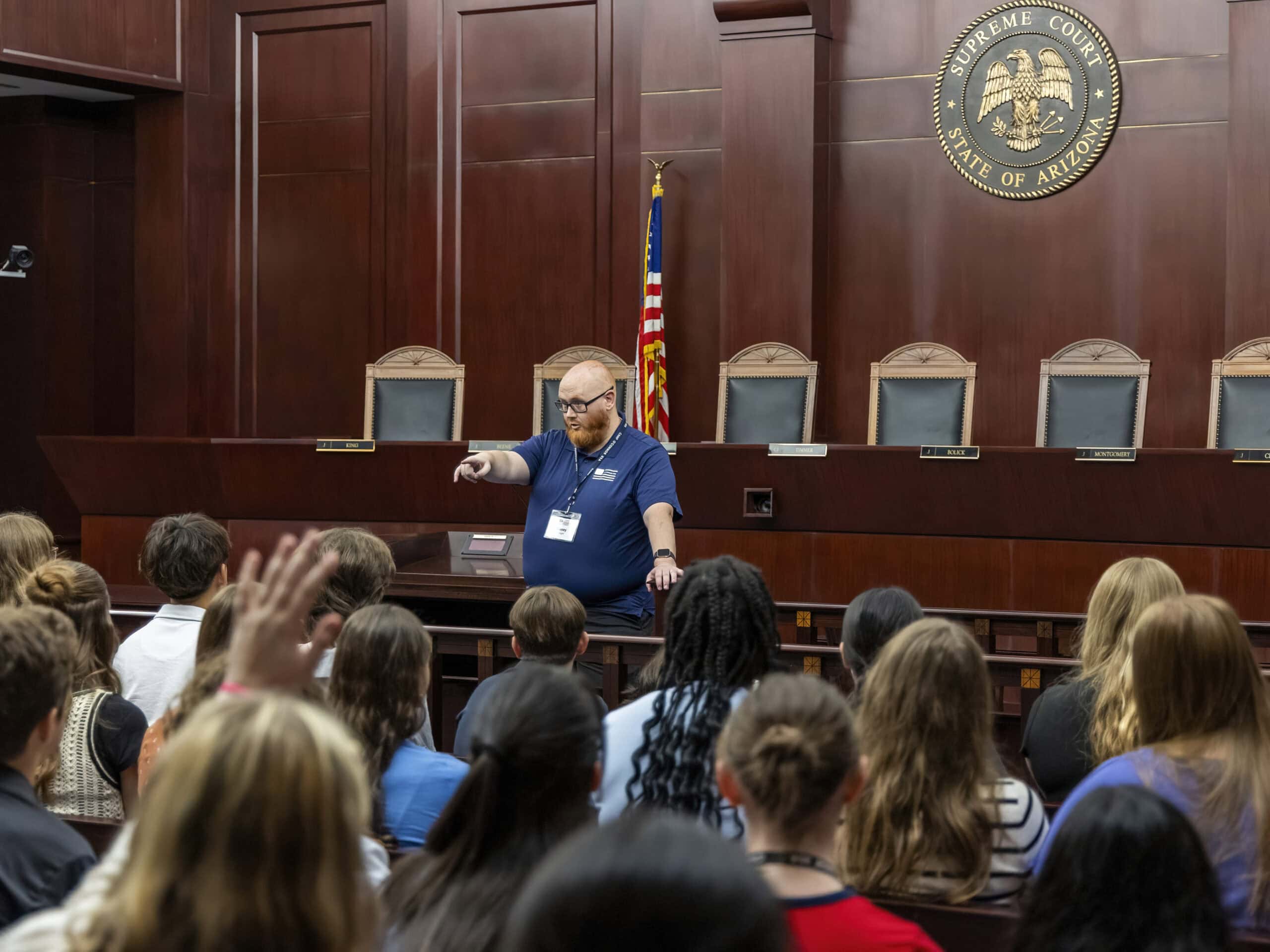 A man stands at the front of a courtroom, speaking to students about the role of a citizen; he points forward while they listen attentively. The room features wooden walls, an American flag, and the Arizona Supreme Court seal.