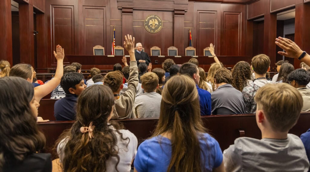 A judge stands at the front of a courtroom speaking to a group of students, many of whom are raising their hands to ask questions—an inspiring scene that highlights Our Impact in fostering legal understanding among youth.