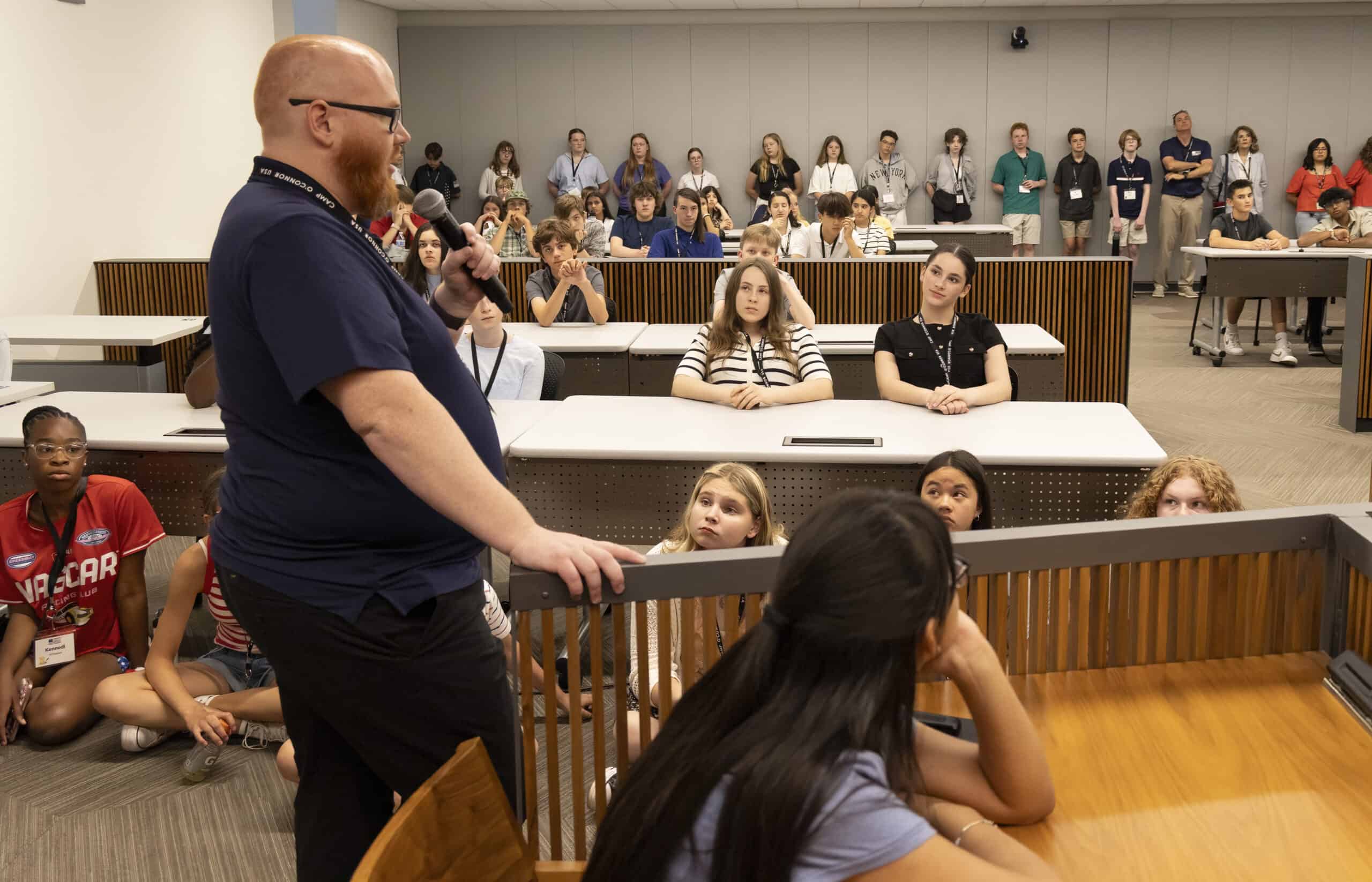 A bearded man speaks to a group of attentive students in a classroom, encouraging each young citizen as some sit at desks and others on the floor, all listening closely.