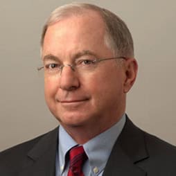 Portrait of an older man with short gray hair and glasses, wearing a suit, light blue shirt, and red tie, set against a plain background.