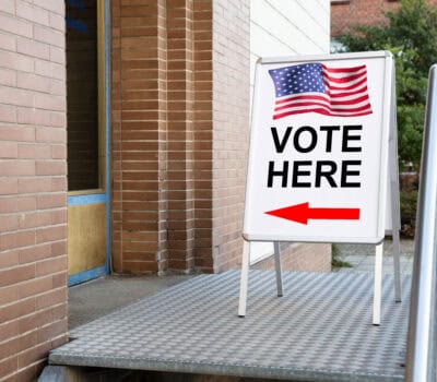 A sign with an American flag and the words "Vote Here" in bold letters, featuring a red arrow pointing left. The sign is on a metal ramp in front of a brick building entrance.