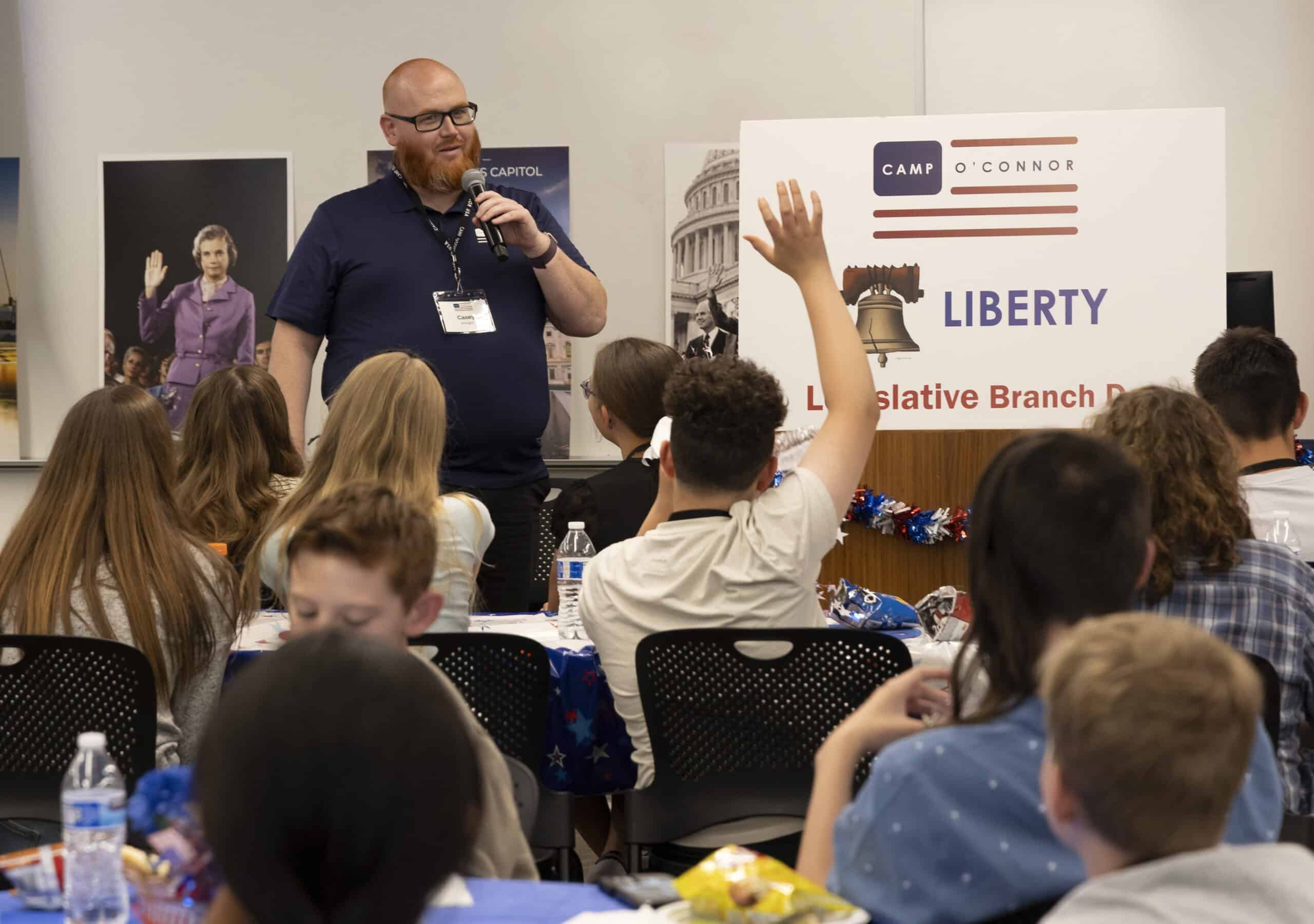 A man speaks into a microphone in front of a group of seated children, one of whom—an eager citizen in the making—has their hand raised. A sign behind him reads “Camp O’Connor Liberty Legislative Branch.”.