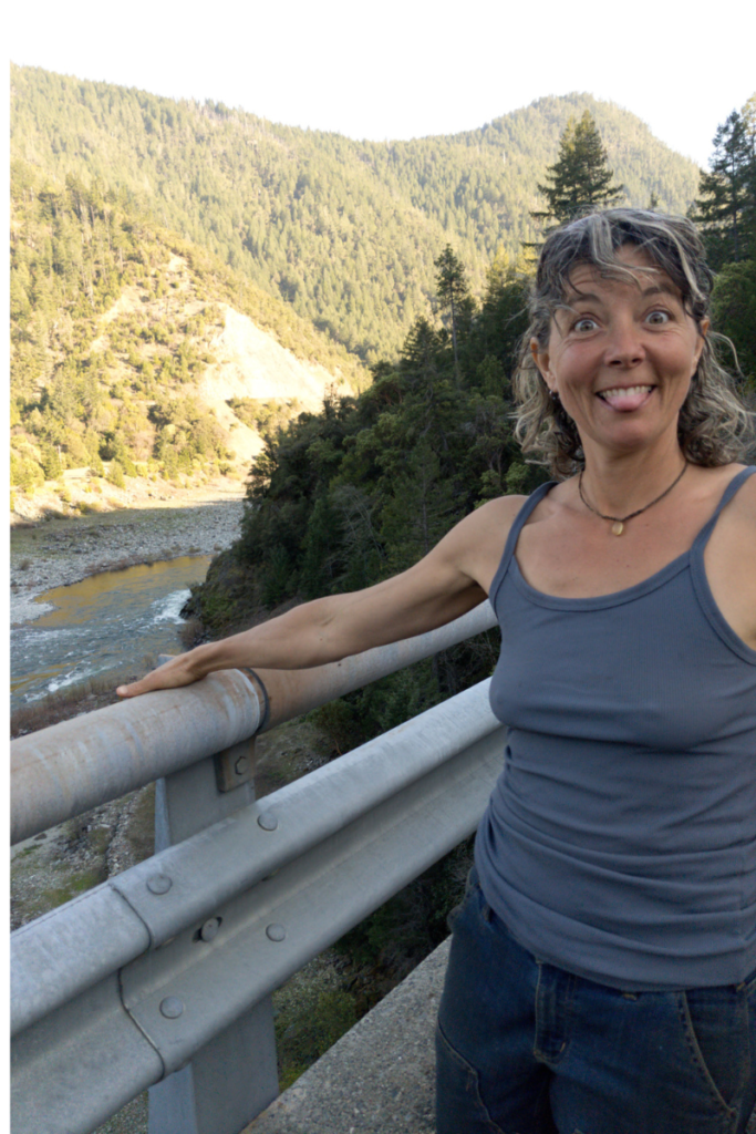 Bobbie smiling playfully and sticking out her tongue while standing on a bridge overlooking a forested mountain valley and river, wearing a gray tank top and jeans, with trees and hills in the background.