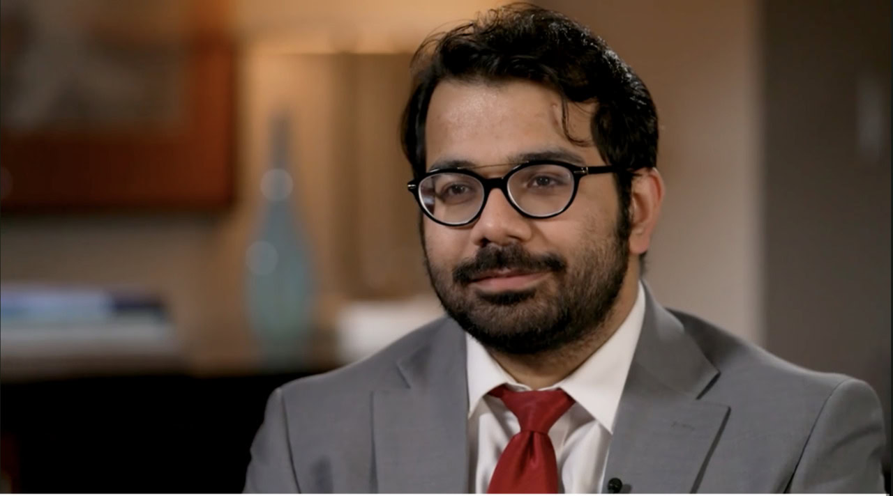 Men in a professional setting, wearing suits and glasses, engaged in a serious conversation or interview.