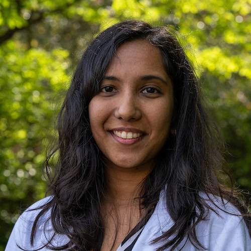 Young woman with dark hair smiling outdoors, surrounded by green foliage, representing health, wellness, and medical professionalism, suitable for cancer treatment or oncological research imagery.