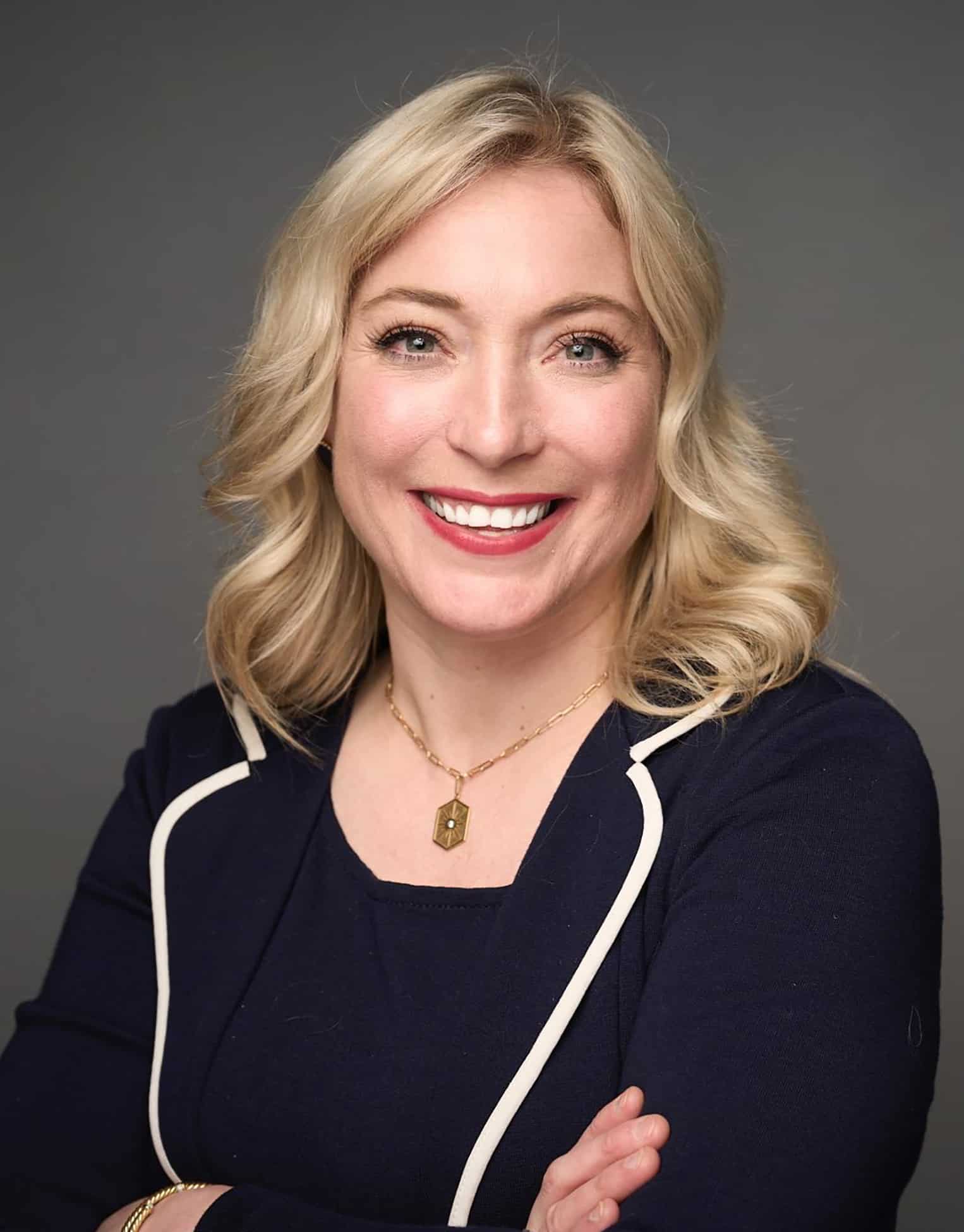 A woman with blonde hair, wearing a navy blazer with white piping, smiling confidently, representing oncology and cancer research professionals, aligned with OncUpdates' focus on advanced cancer insights.