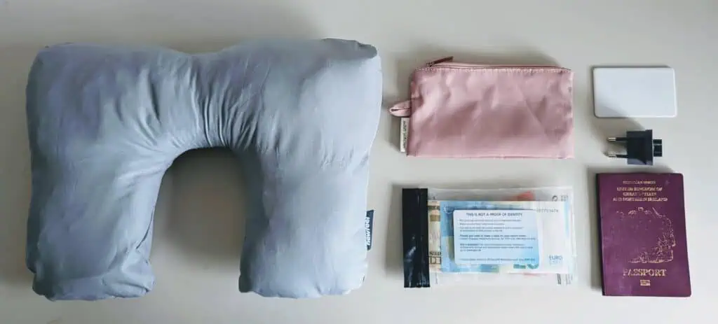 Flat lay showing international travel essentials including passport, pillow case stuffed with jacket, GHIC card, chipolo card, plug adapter, and pink zip pouch
