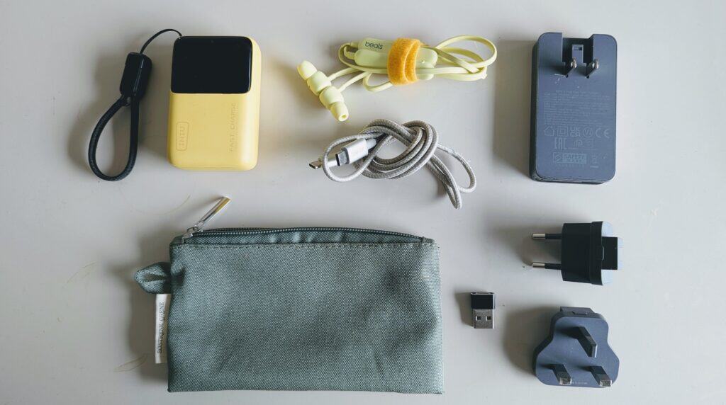 Flat lay of lightweight tech kit including power bank, Beats earbuds, charging cable, plug with interchangeable ends, USB adapter, and a green tech pouch