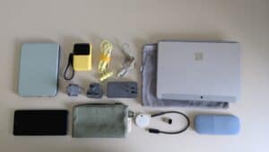 Flat lay of a minimalist travel tech kit, including a Microsoft Surface tablet with sleeve, Microsoft Arc Mouse, compact power bank, wired earbuds, USB-C cables, international plug adapters, universal USB charger, Chipolo tracker, smartphone, zipped tech pouch, and Kobo e-reader in a green case. Lightweight, colour-coordinated setup for carry-on-only digital nomads.