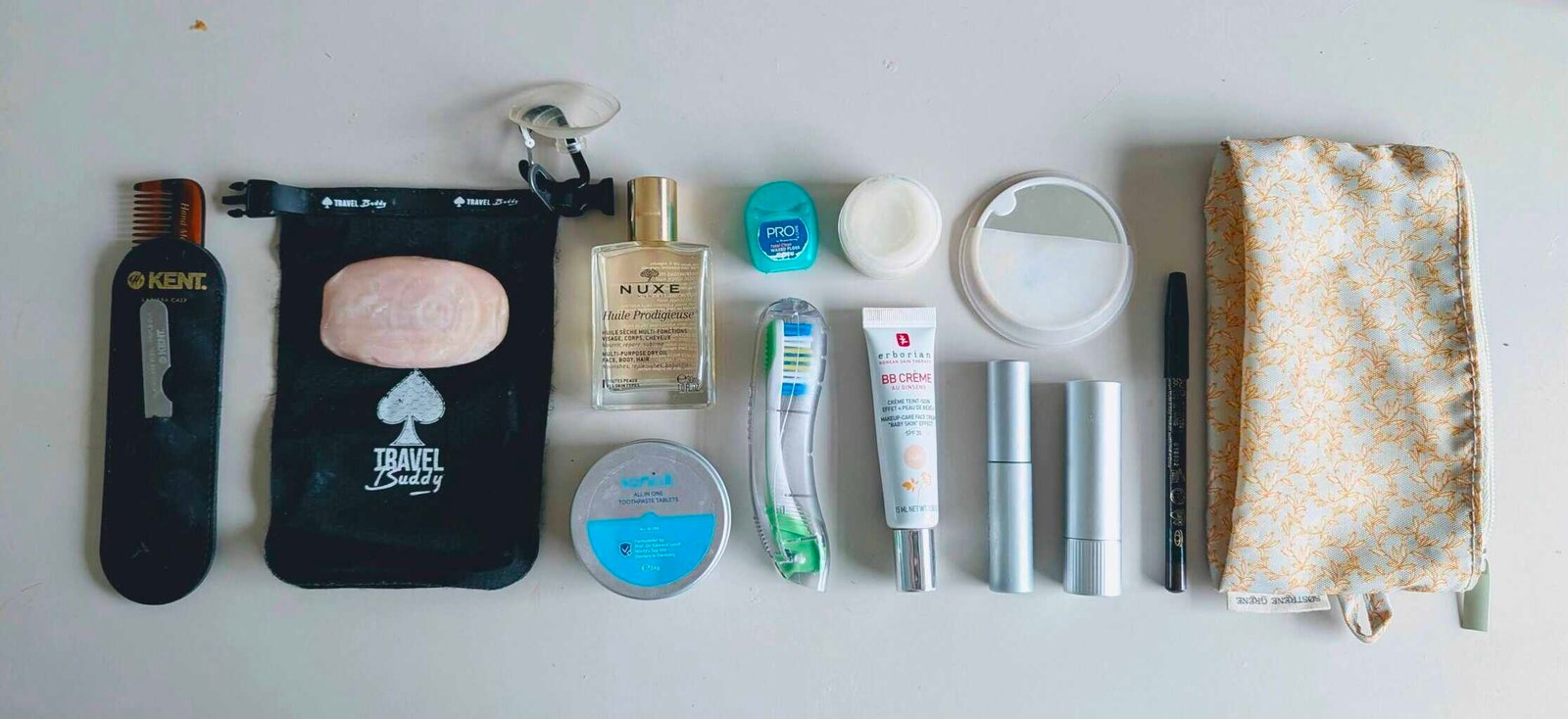 Flat lay of minimalist carry-on toiletries for travel, including folding toothbrush, toothpaste tablets, shampoo bar, multi-use oil, BB cream, solid deodorant, compact comb, and a small floral pouch. Lightweight setup from One Small Bag.