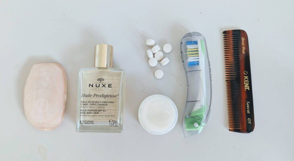 Minimalist travel toiletries laid out in a row — solid soap, dry oil, toothpaste tablets, moisturiser, folding toothbrush, and a compact comb — covering the essentials without excess.