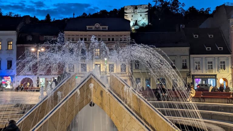 Council Square Brasov at night with a view of the fountain and the White Tower and Carpathian Mountains in the background
