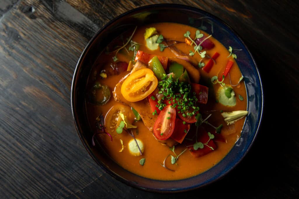 A bowl of colorful soup with various vegetables and garnishes, presented artistically on a dark wooden surface.