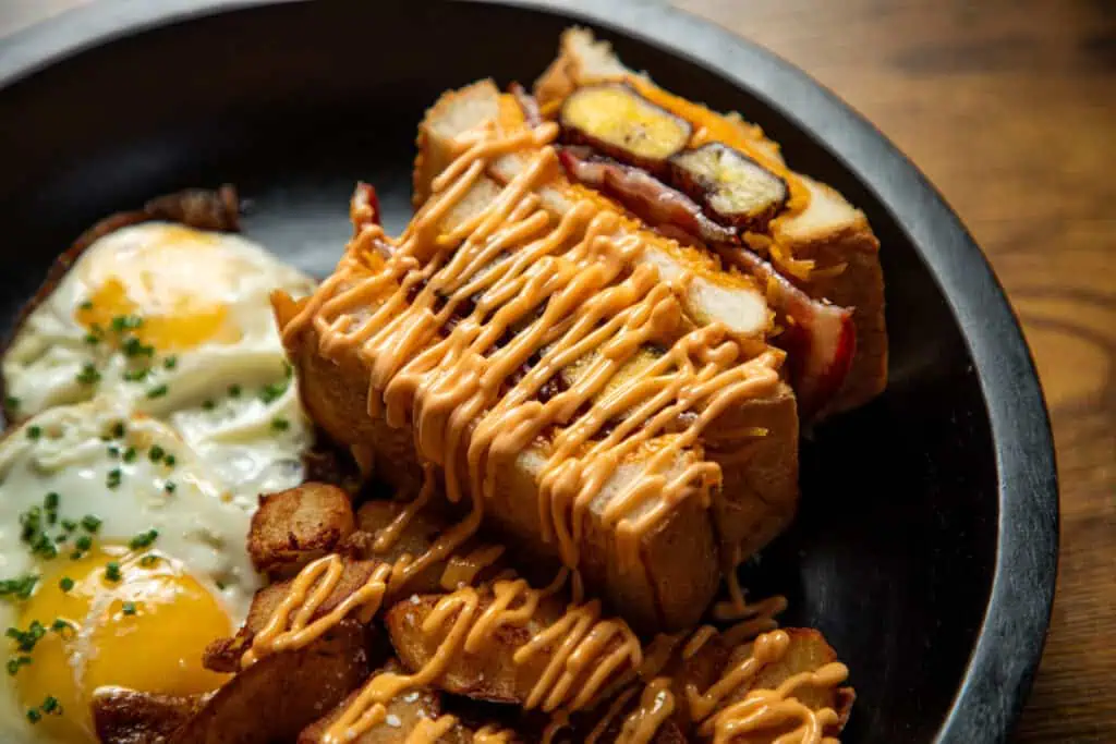 A plate featuring a sandwich with toppings, paired with two fried eggs and seasoned potato wedges.