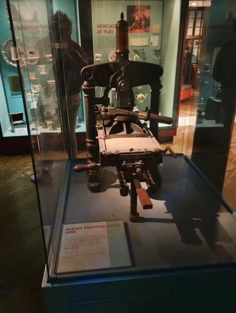 Close-up of an antique Albion printing press from 1868, showcased in a glass case at a museum. The device is made of dark metal and wood, highlighting early printing technology used in traditional print shops.