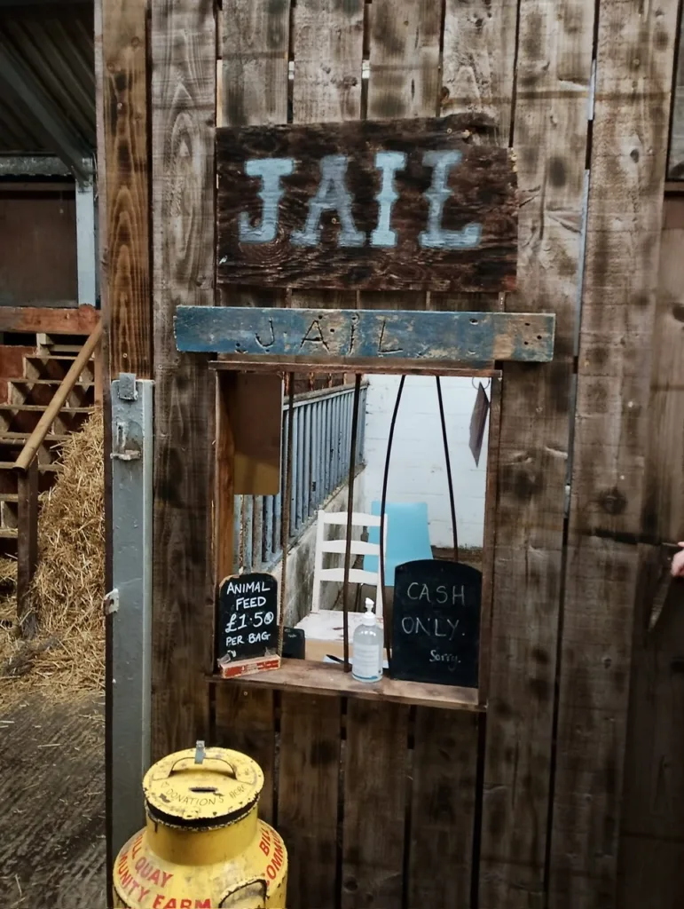 Rustic wooden jail sign at animal farm entrance with "JAIL" painted in blue, featuring a small window and a "Cash Only" sign, creating a quirky farm-themed attraction.