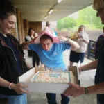 Group of friends celebrating a birthday with a cake and candles outdoors, with a young girl wearing a pink party hat and others smiling and enjoying the occasion.