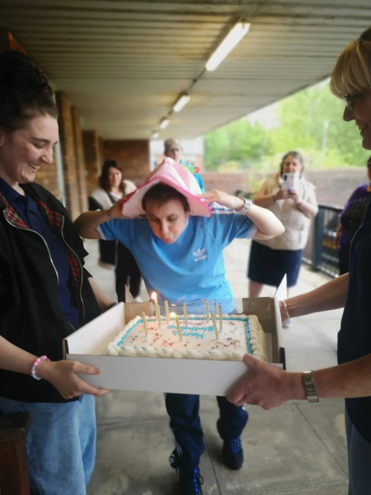 Group of friends celebrating a birthday with a cake and candles outdoors, with a young girl wearing a pink party hat and others smiling and enjoying the occasion.