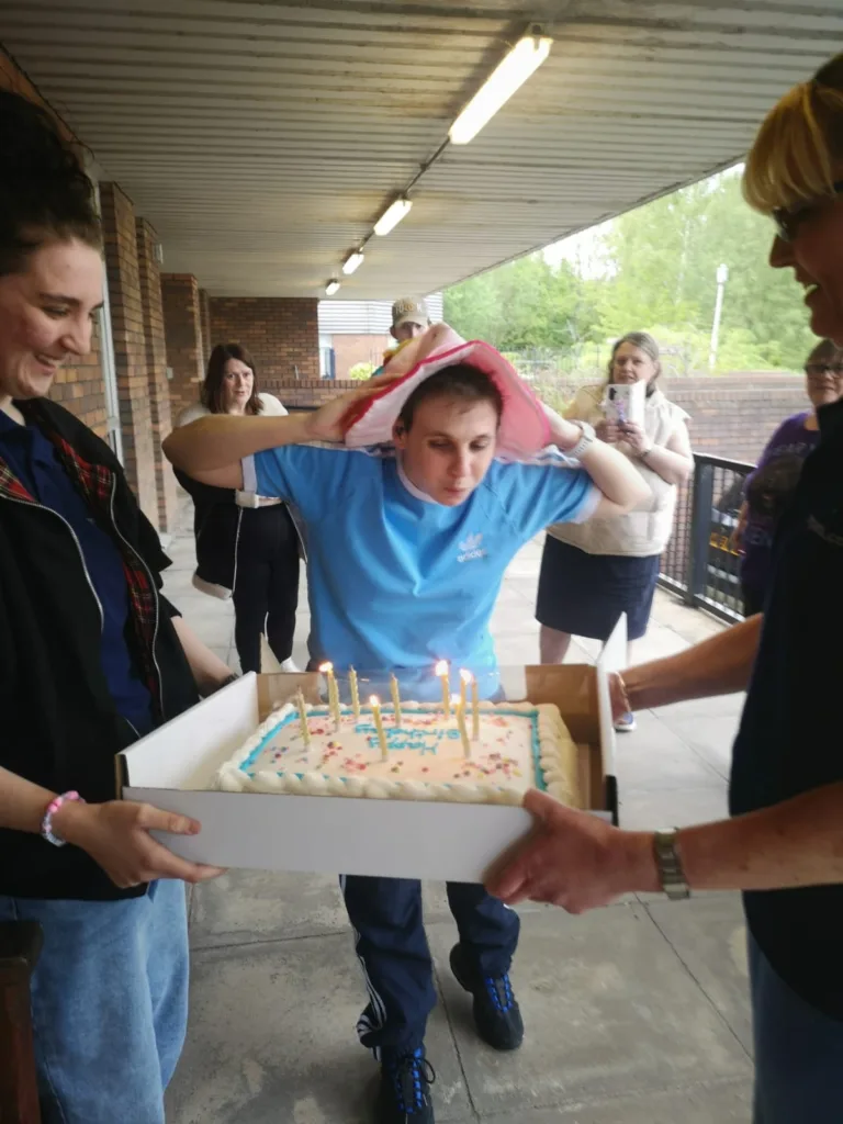 Young girl with a birthday cake and candles, surrounded by friends and teachers outdoors at school.
