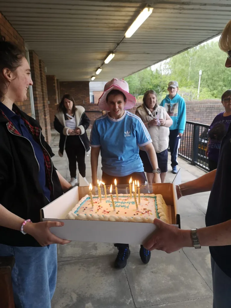 A group of people celebrating a birthday with a cake outdoors at Original Support Ltd. The scene features a woman in a pink hat and a cake with lit candles, surrounded by friends in a casual setting.