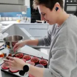 Young person with short hair, wearing a grey sweatshirt and a smartwatch, placing cupcakes in a tray.