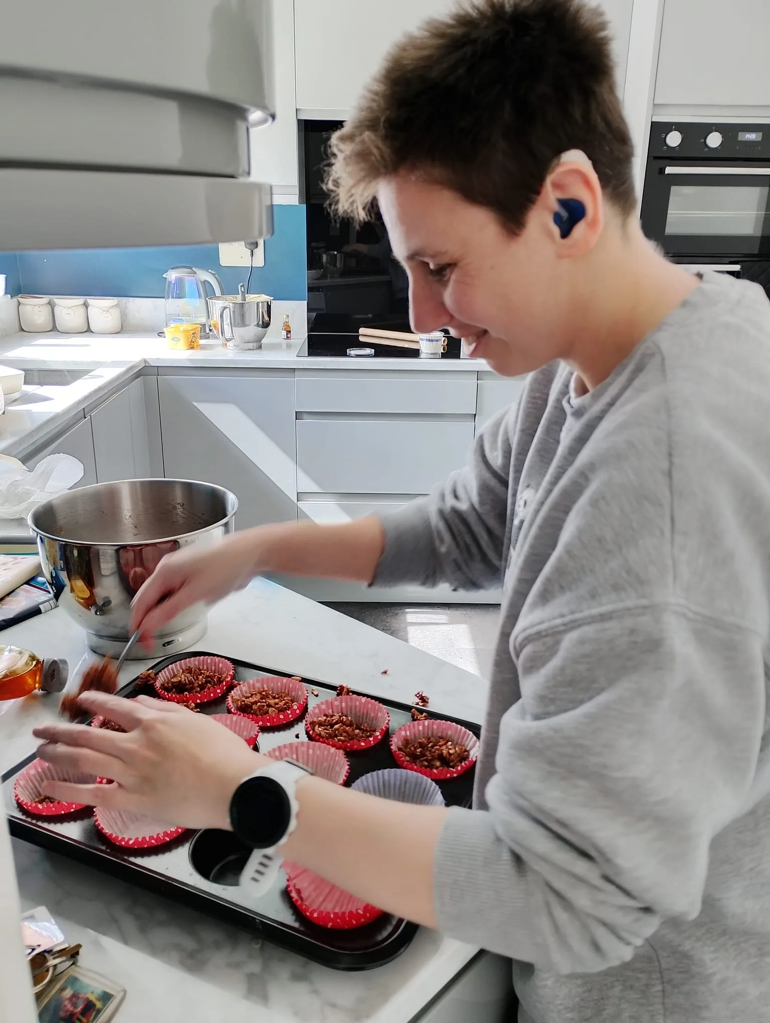 Young person with short hair, wearing a grey sweatshirt and a smartwatch, placing cupcakes in a tray.