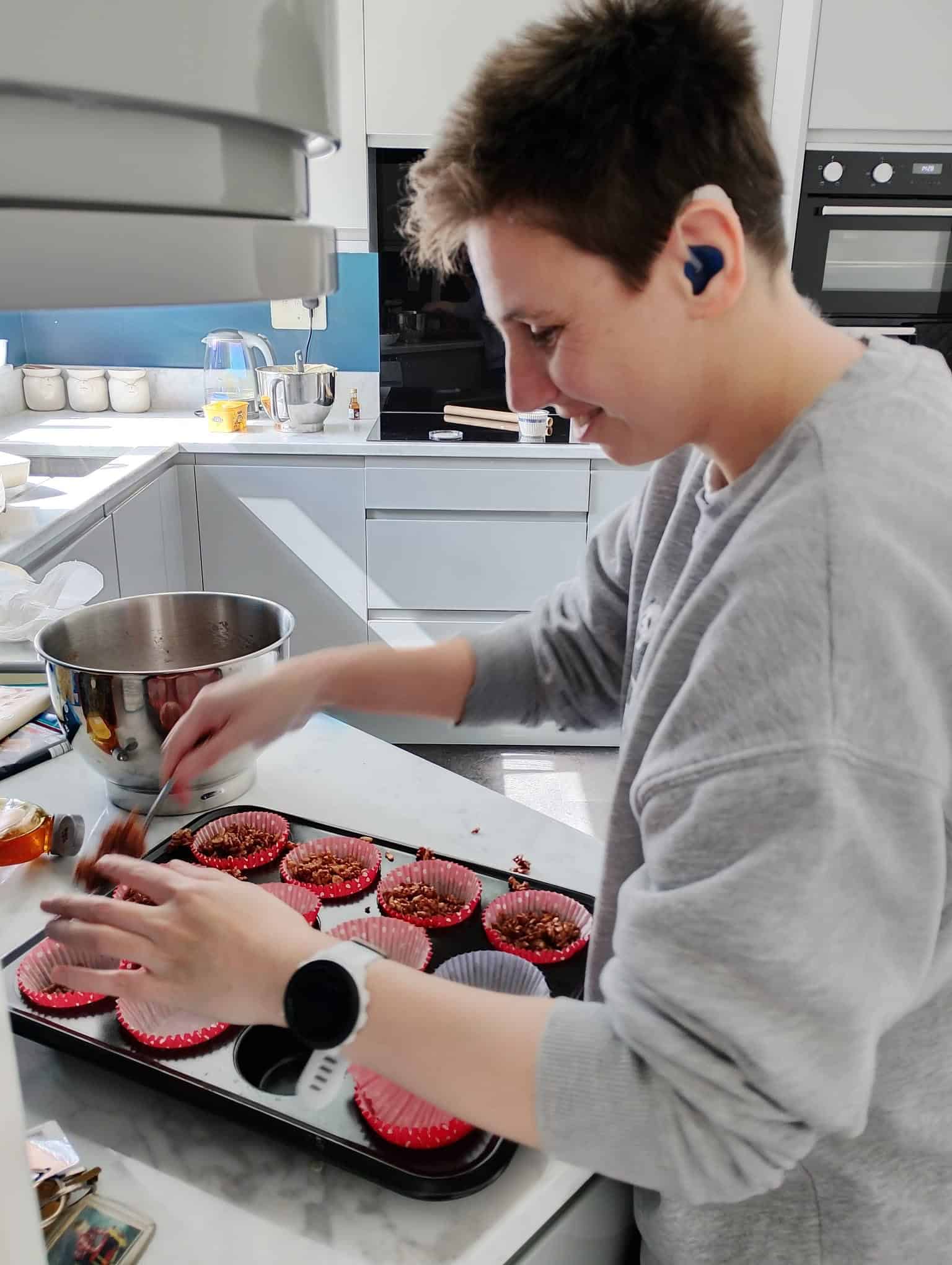 Young person with short hair, wearing a grey sweatshirt and a smartwatch, placing cupcakes in a tray.
