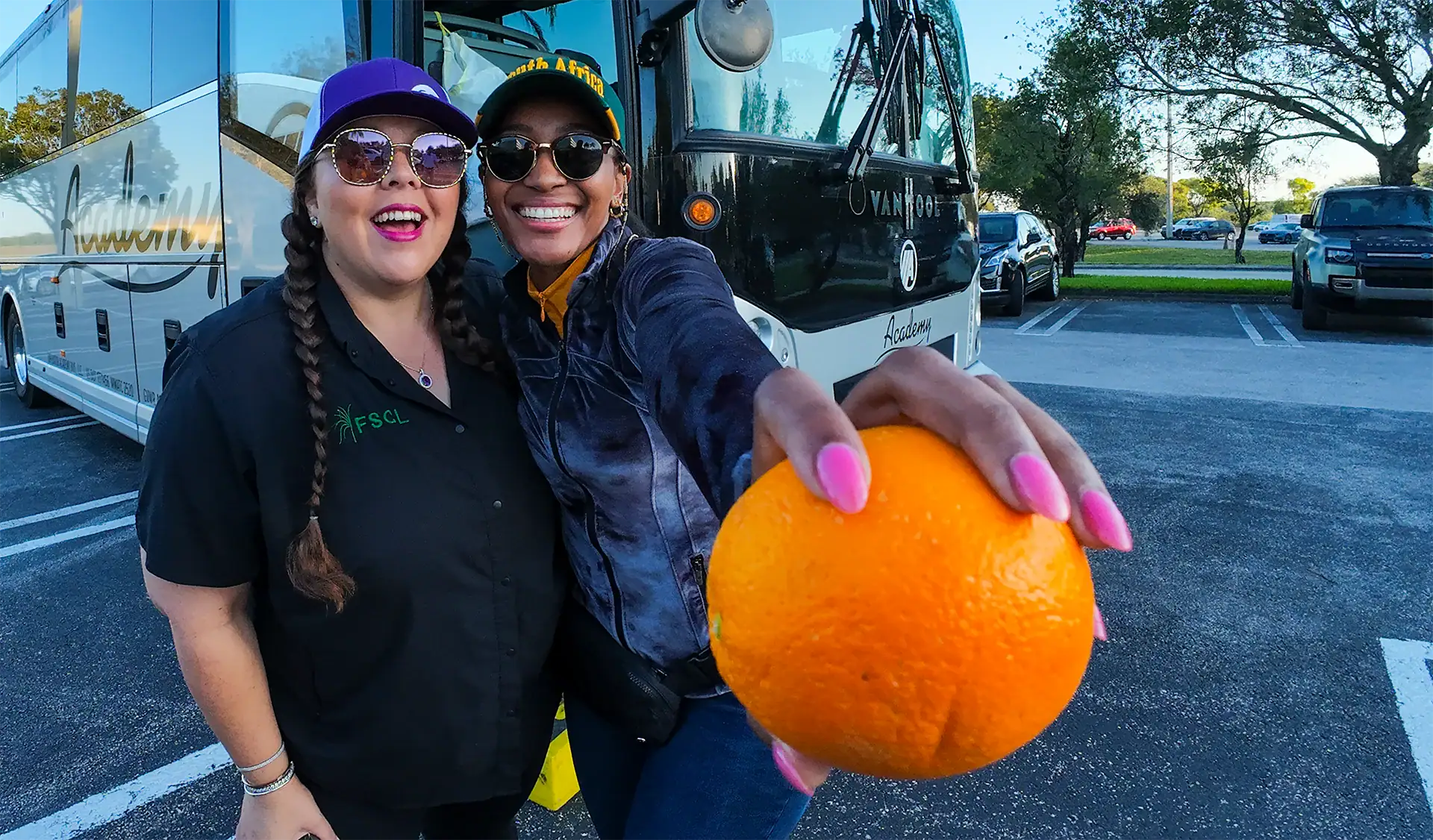 Women smiling, wearing sunglasses, and casual clothing, with a bus in the background.