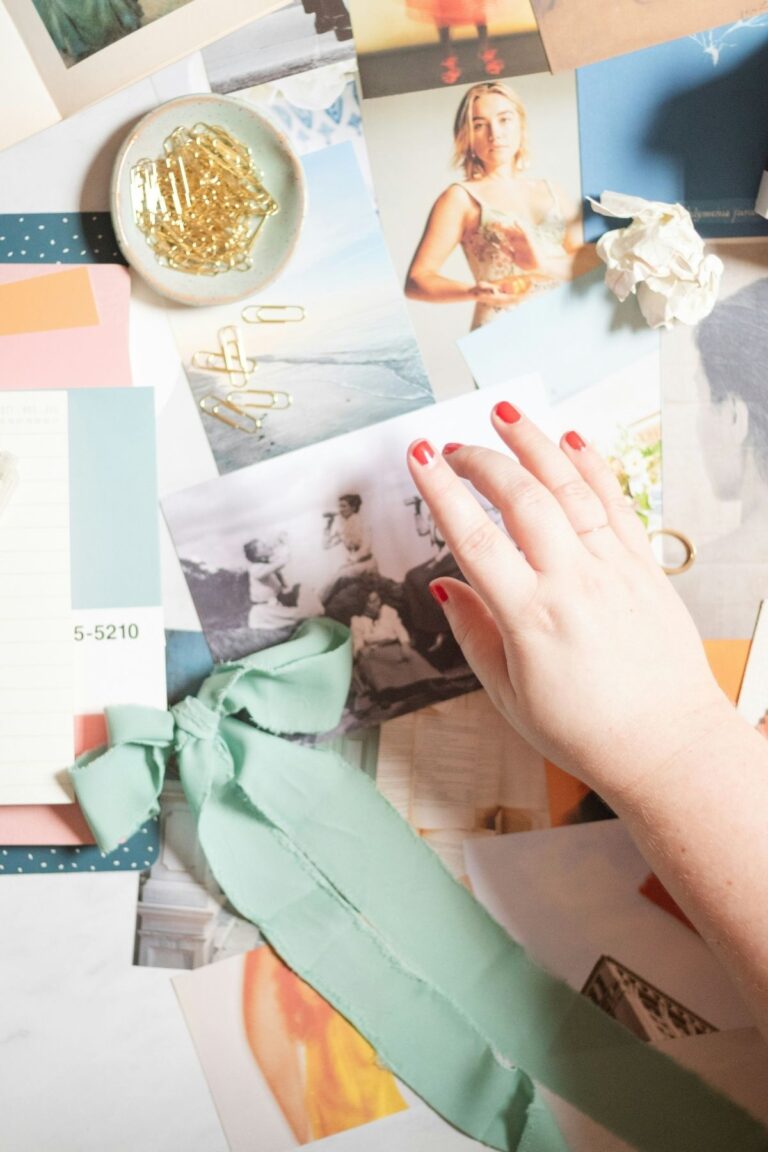 Flay lay of a desk covered by numerous overlapping photographs. Also on the table is a pale green fabric bow and matching circular dish containing gold paperclips. A white woman's hand with red painted nails is reaching for a photograph on the table.