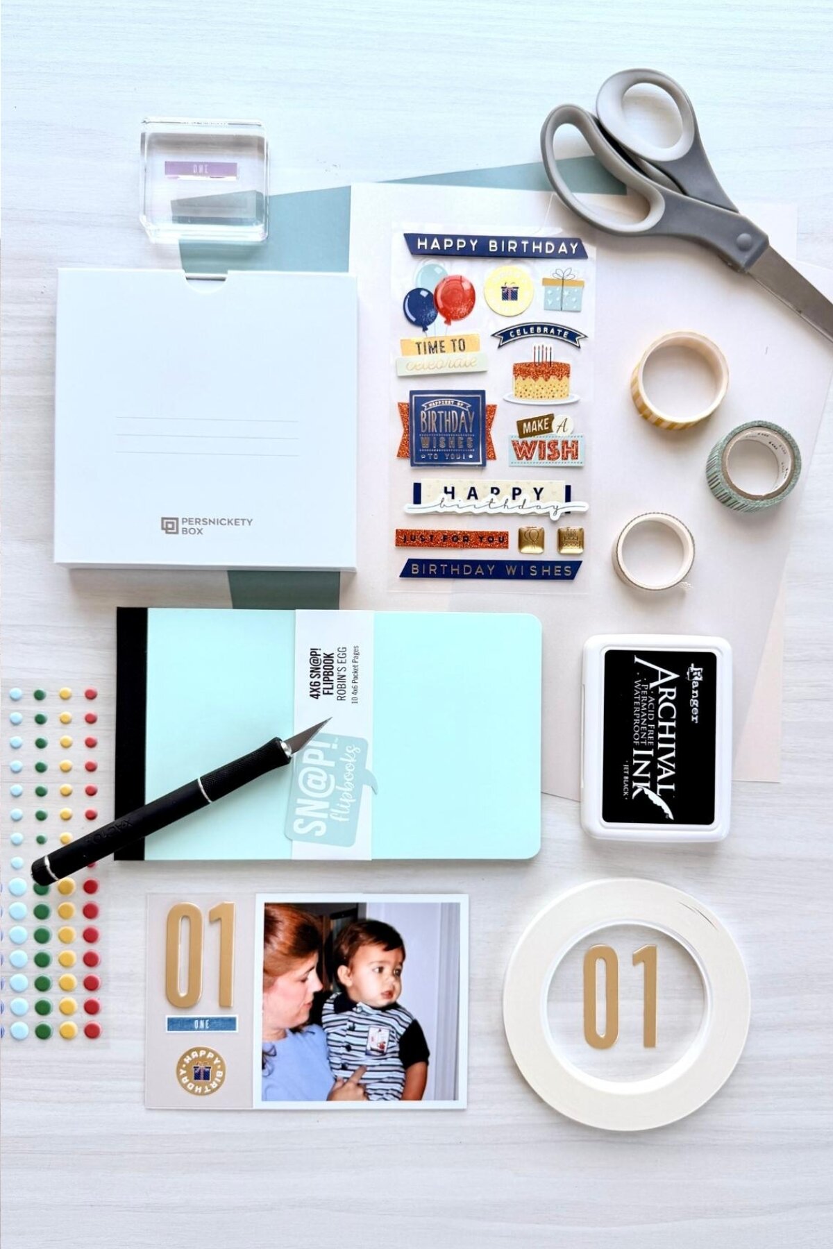 Flatlay of scrapbooking supplies needed to create the mini album. Supplies include from top to bottom: stamp block, scissors, Persnickety Box, stickers, washi tape, mini album, ink pad, embellishments, tape, and die cut numbers. A sample page from the completed album is also shown.