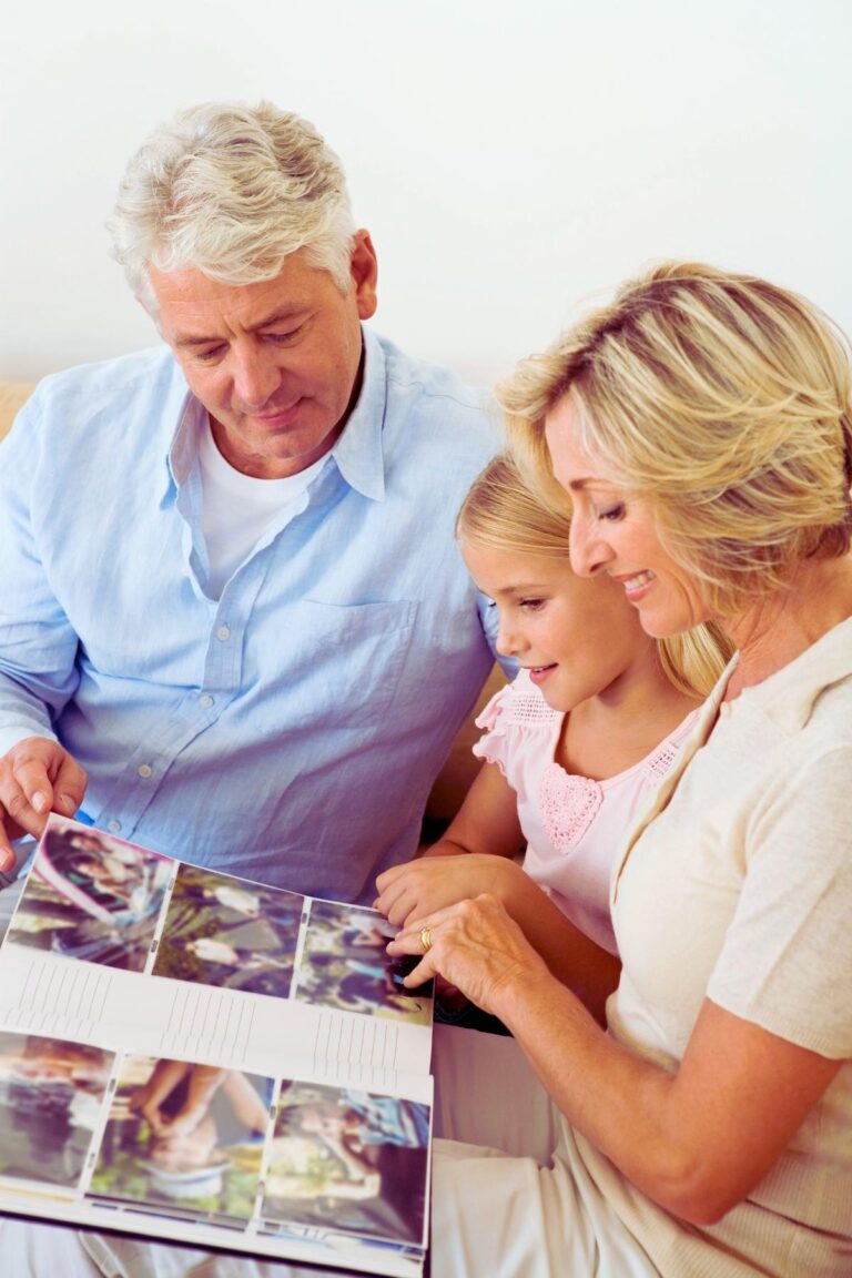 Grandparents and their granddaughter are sitting down looking at a photo album together. The granddaughter is about six years old and is seated between her grandparents. The photo album is setting on their laps.