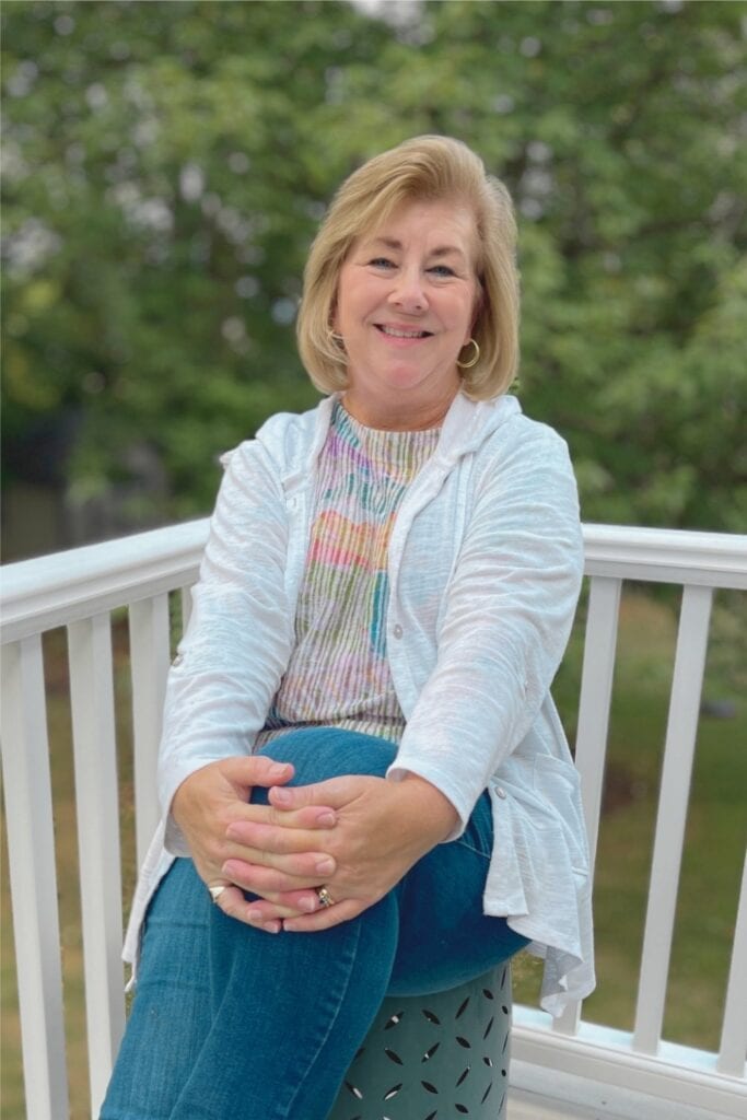 Casual photo of Christy, owner of Paper Hideaway, sitting outdoors with a white porch railing and green trees behind her.