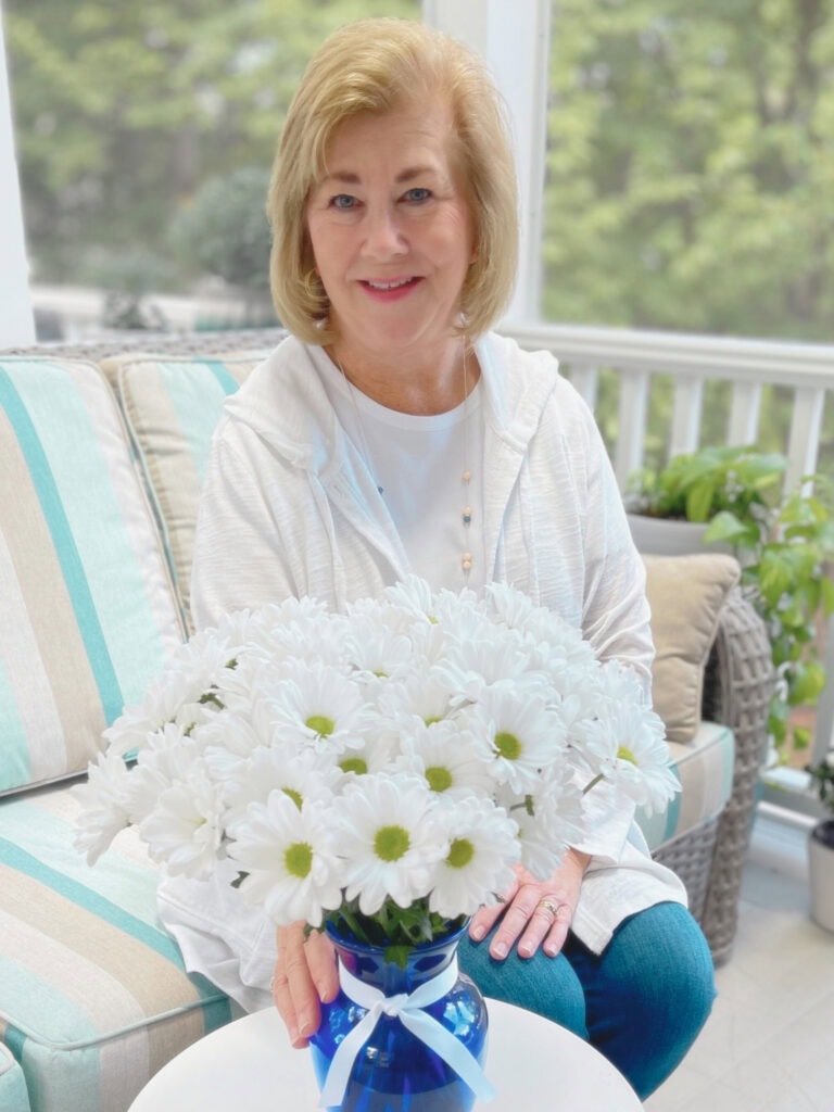 Christy smiling and sitting outdoors on a porch with a blue vase filled with white daisies in front of her.