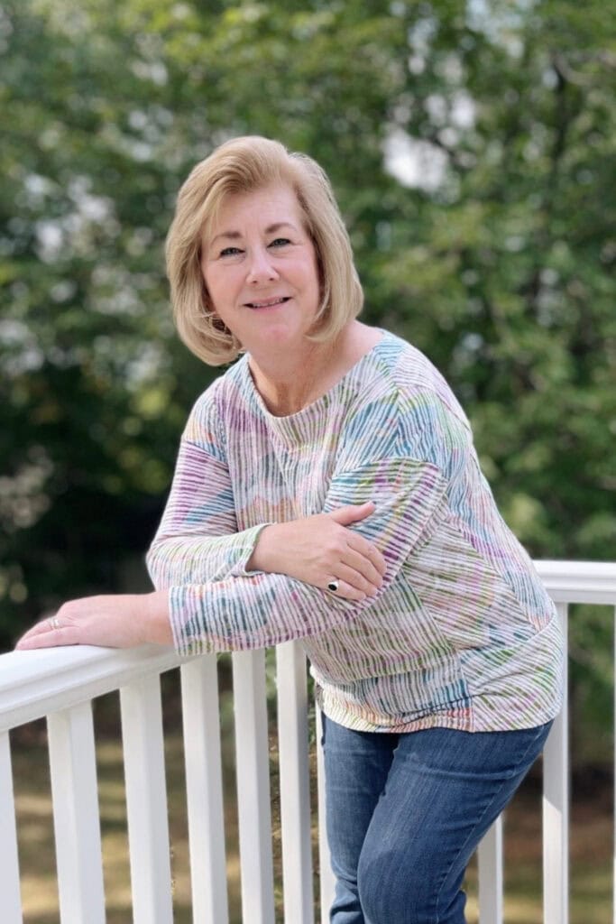 Photo of Christy, owner of Paper Hideaway, taken outdoors. She is middle-aged, caucasian and has shoulder length light brown hair. Christy is leaning on a white porch railing and looking at the camera. She is dressed in blue jeans and a pastel printed shirt.