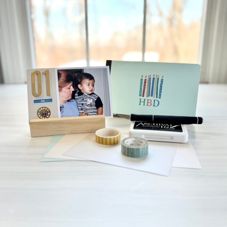 Photo of a mini-scrapbook album setting on a white desk surrounded by an ink pad, washi tape and x-acto knife. The album is a honeydew color and is decorated with a birthday sticker. The sticker shows birthday candles with the letters HBD beneath the image. To the left of the album is a completed 4"x6" page from the album using a 4"x4" Persnickety Box print.