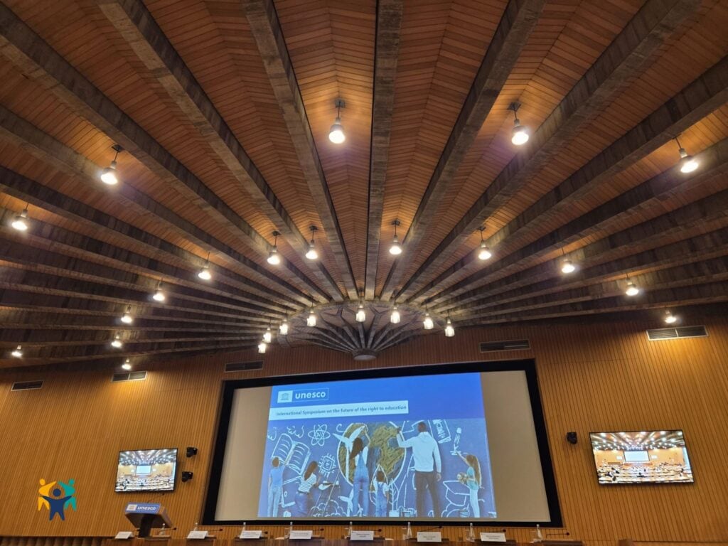 Photo of a UNESCO conference hall with a dramatic wooden ceiling and a large screen showing the “International Symposium on the future of the right to education,” with empty panel seats and side screens displaying the same image.