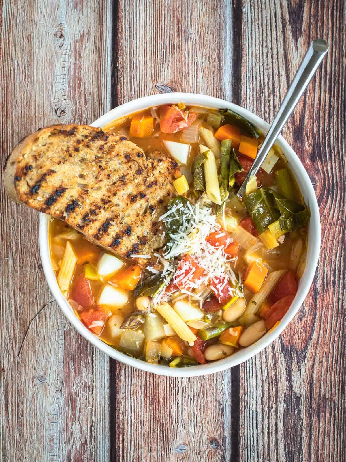 a bowl of minestrone soup with grilled bread, shredded parmesan cheese, and a spoon