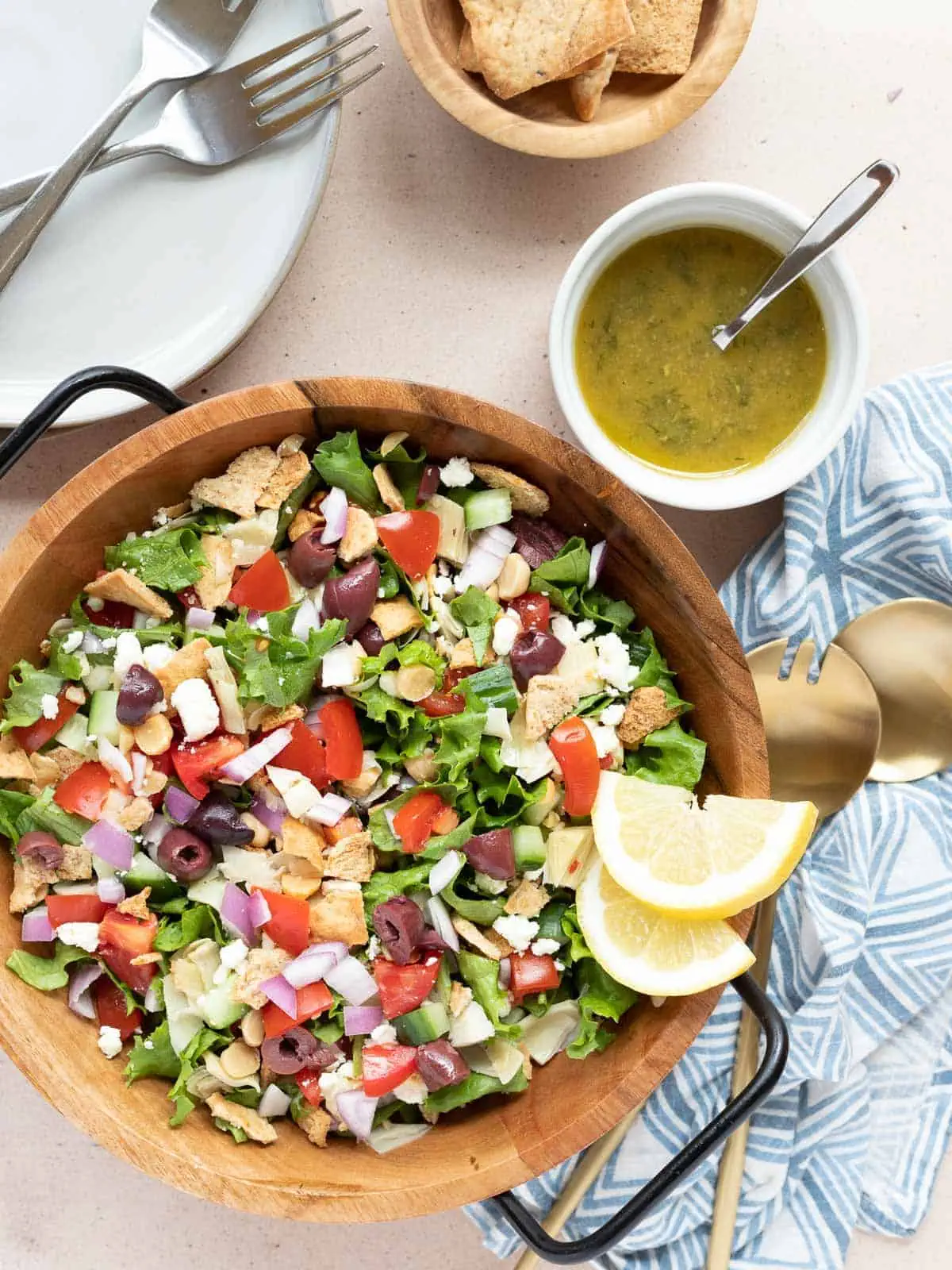 Loaded Greek Salad in a bowl. Garnished with lemons. Surrounded by a bowl of dressing, a bowl of pita chips, a cloth napkin, and salad servers.