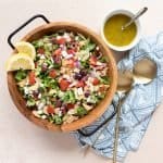 Loaded Greek Salad in a bowl garnished with lemon slices. Sitting next to the bowl is a ramekin of dressing with a spoon, a cloth napkin, and salad servers.