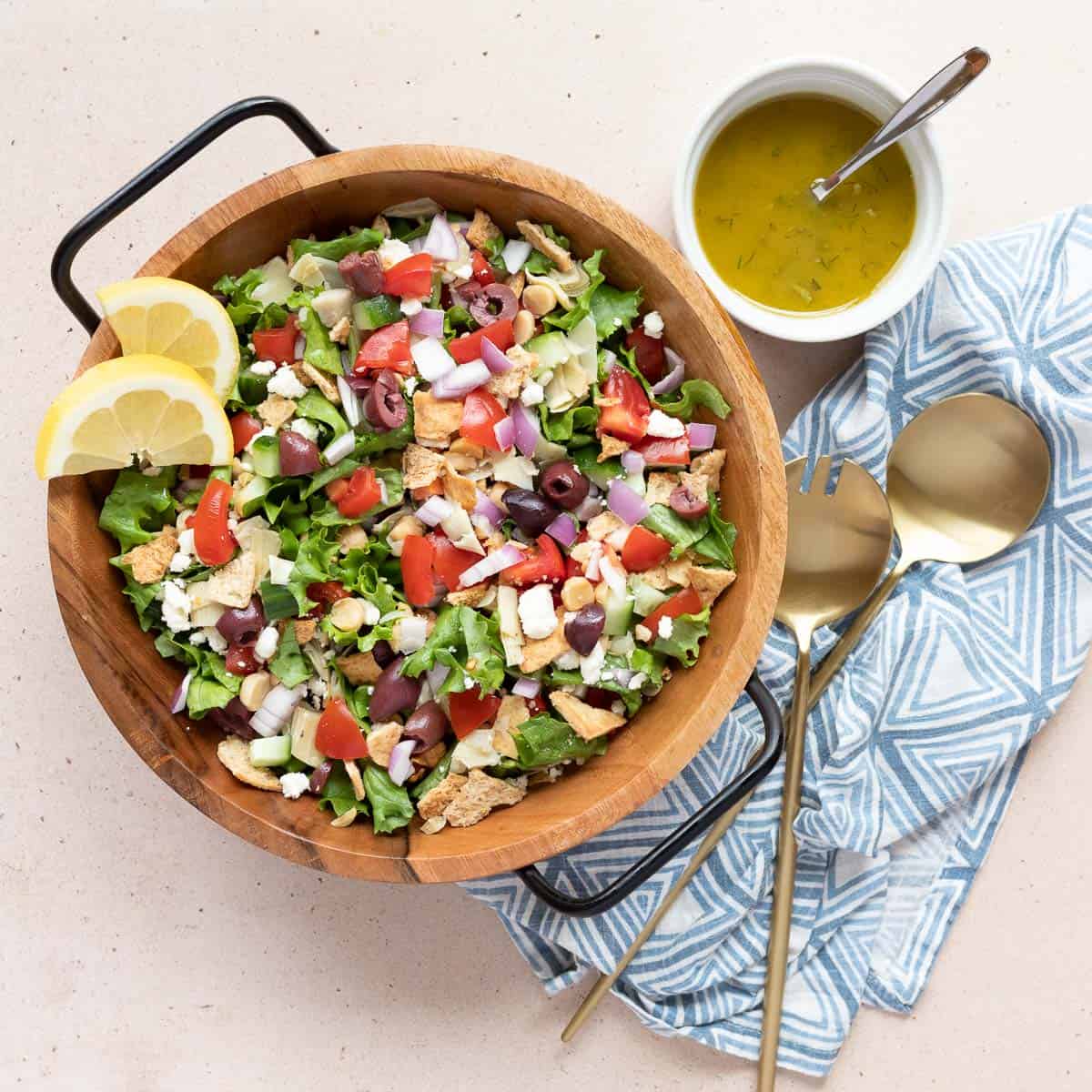 Loaded Greek Salad in a bowl garnished with lemon slices. Sitting next to the bowl is a ramekin of dressing with a spoon, a cloth napkin, and salad servers.