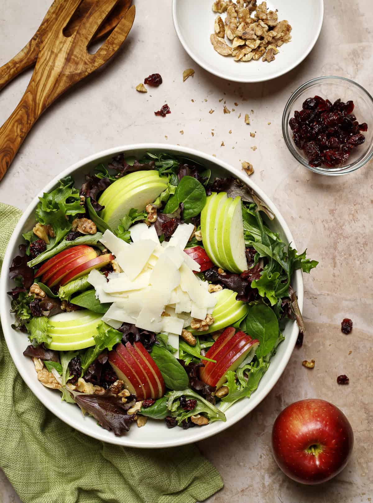 Apple and Asiago Salad features crisp salad greens, crunchy apples, sweet-tart dried cranberries, and toasted walnuts. It is dressed with maple-Walnut vinaigrette. Extra walnuts and cranberries sit nearby.