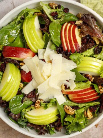 Apple and asiago salad in a bowl, featuring crisp salad greens, sliced red and green apples, shaved asiago cheese, toasted walnuts, and tart dried cranberries. It is dressed with an easy maple-Dijon vinaigrette.