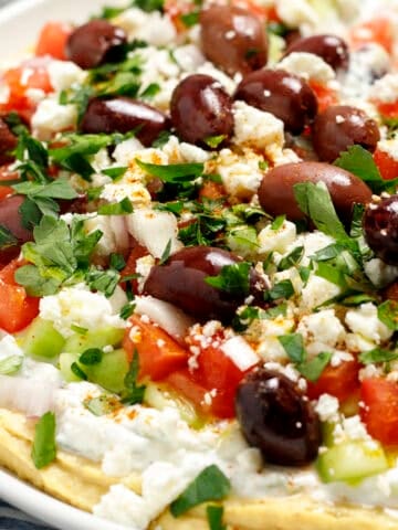 A plate filled with Greek layer dip including hummus, tzatziki, cucumbers, tomatoes, olives, shallot, and parsley.