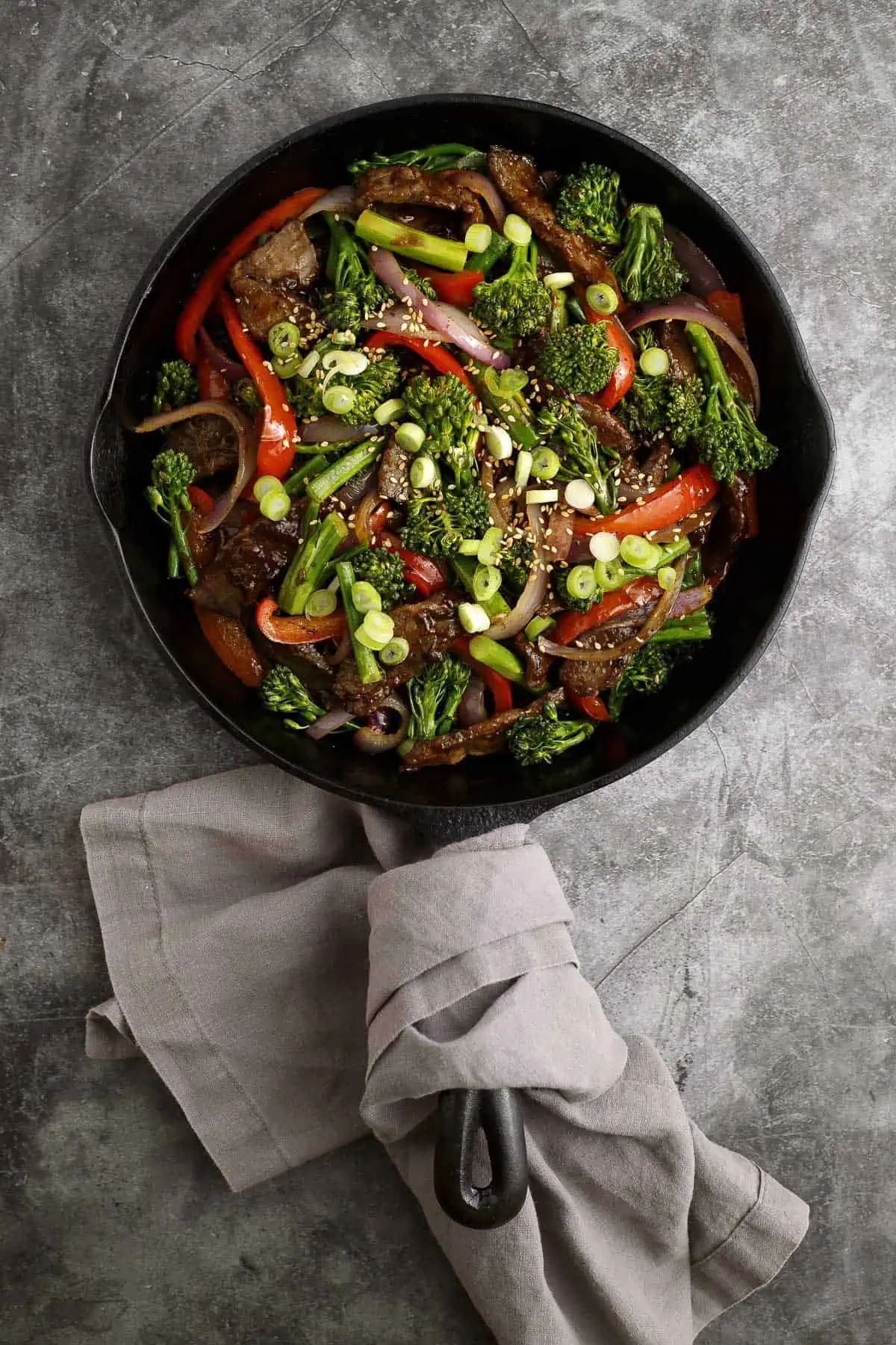this photo contains a cast iron pan filled with beef and broccolini stir fry containing red bell peppers and red onions, garnished with toasted sesame seeds and sliced scallions. a napkin is wrapped around the handle.