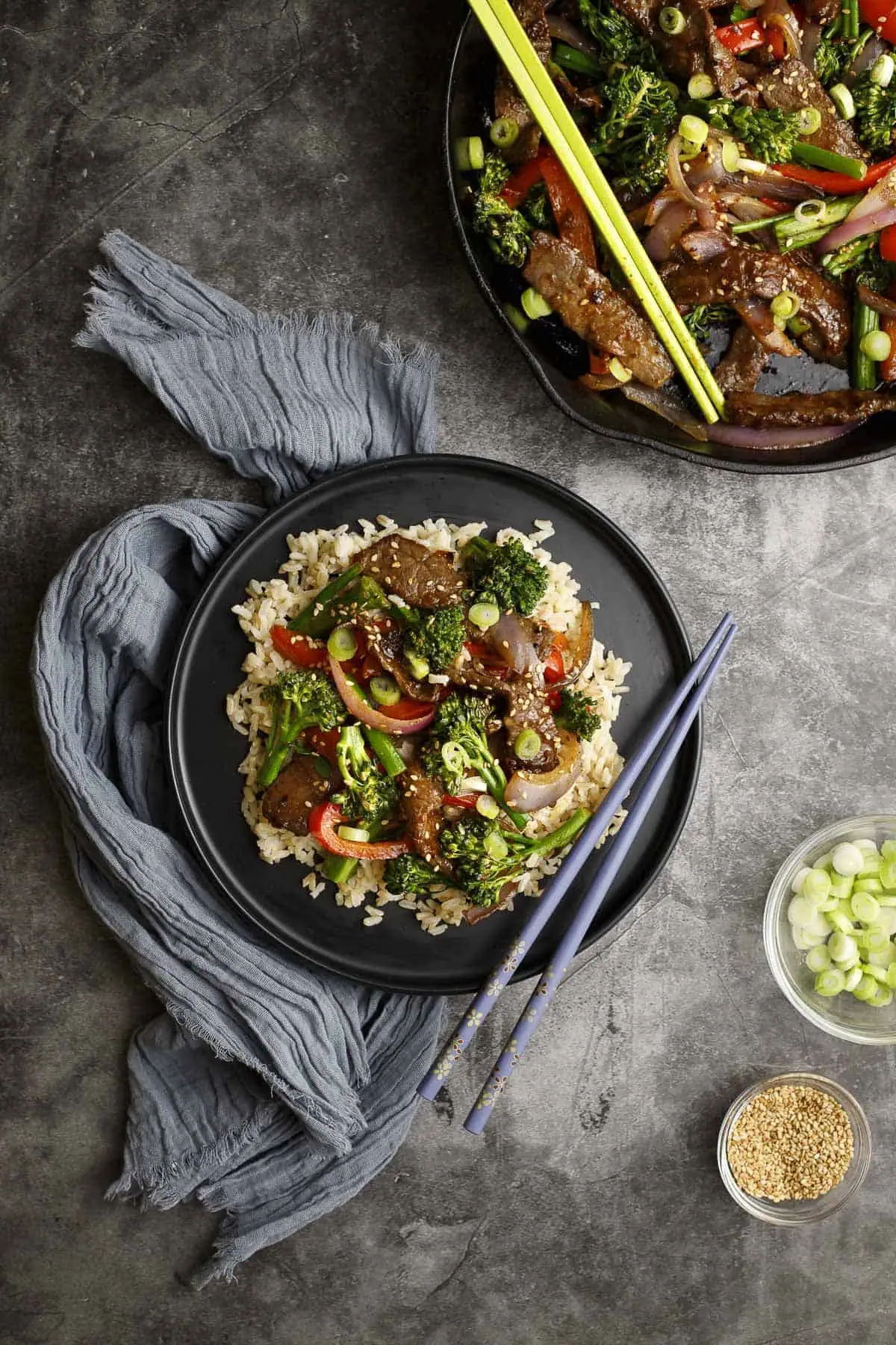 Beef and broccolini stir fry is shown on a plate with chopsticks. A bowl of toasted sesame seeds and one of sliced scallions sits nearby.
