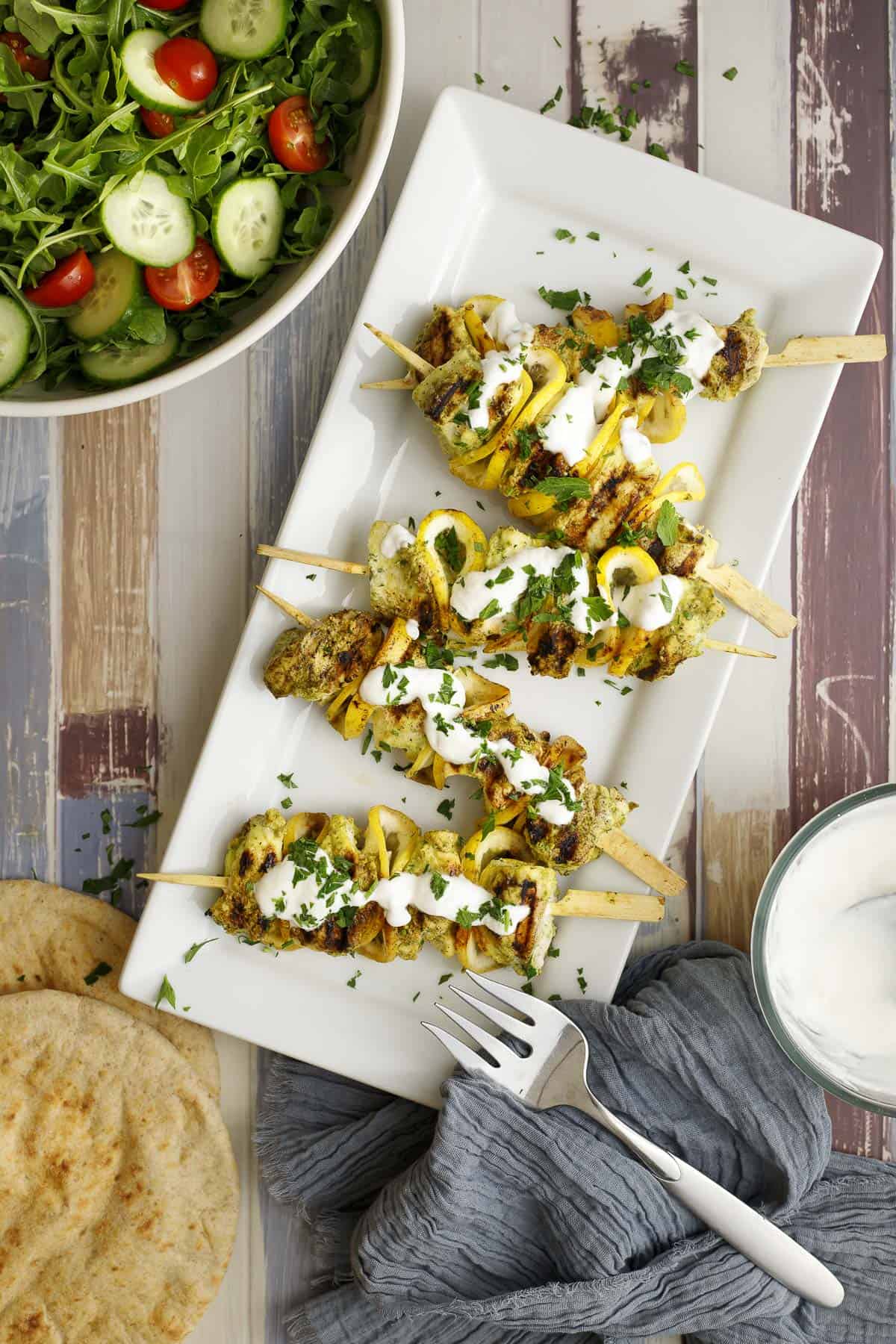 easy grilled halibut skewers on a platter topped with lemon garlic yogurt sauce and surrounded by a salad, pita bread, and extra sauce