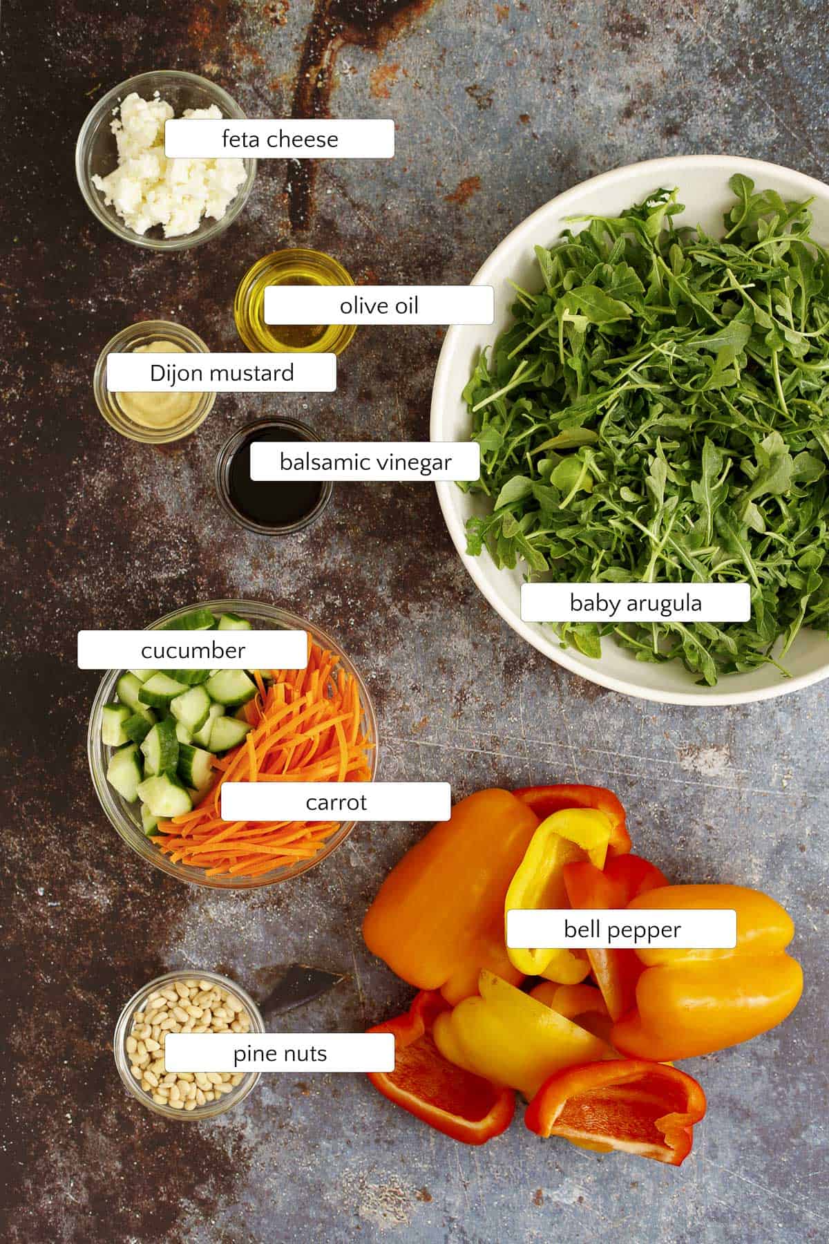 ingredients for grilled pepper salad are arugula, bell peppers, cucumbers, carrots, pine nuts, feta cheese, balsamic vinegar, Dijon mustard, olive oil, salt, and pepper.