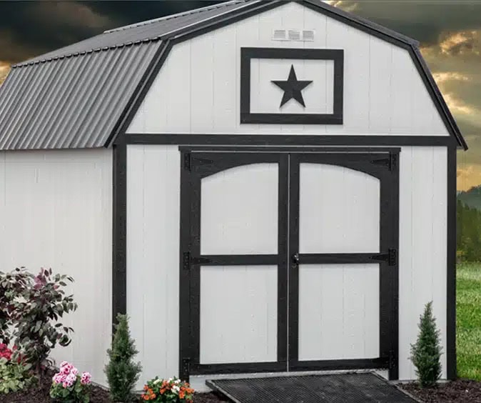 Gemini said A white, gambrel-roofed storage shed stands in a landscaped yard under a dramatic, cloudy sky. The shed features a dark gray corrugated metal roof and striking black trim that outlines the double doors and the roofline. A prominent black star is mounted within a rectangular black frame on the front gable. Small evergreen shrubs and colorful flowers are planted at the base, and a black ramp leads up to the arched double doors.