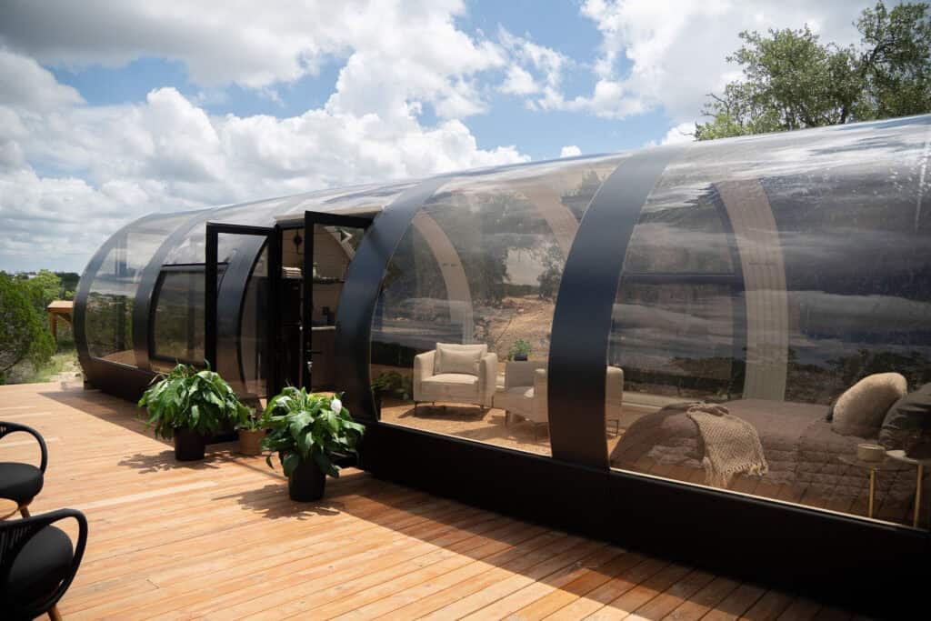 Sleek, modern transparent glamping pod with interior seating and bedroom, outdoor wooden deck with potted plants, under a bright sky, ideal for luxury outdoor lodging or unique travel experiences.